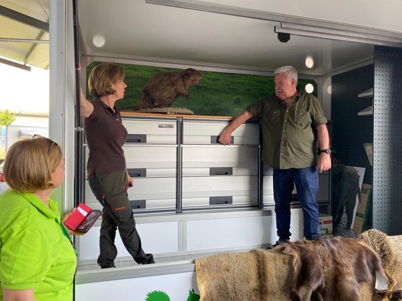 Ein ausgestopfter Biber steht auf einem Tisch, beobachtet von drei Personen in Freizeitkleidung. Der Raum hat Metallregale und eine grüne Hintergrundszene mit Gras.